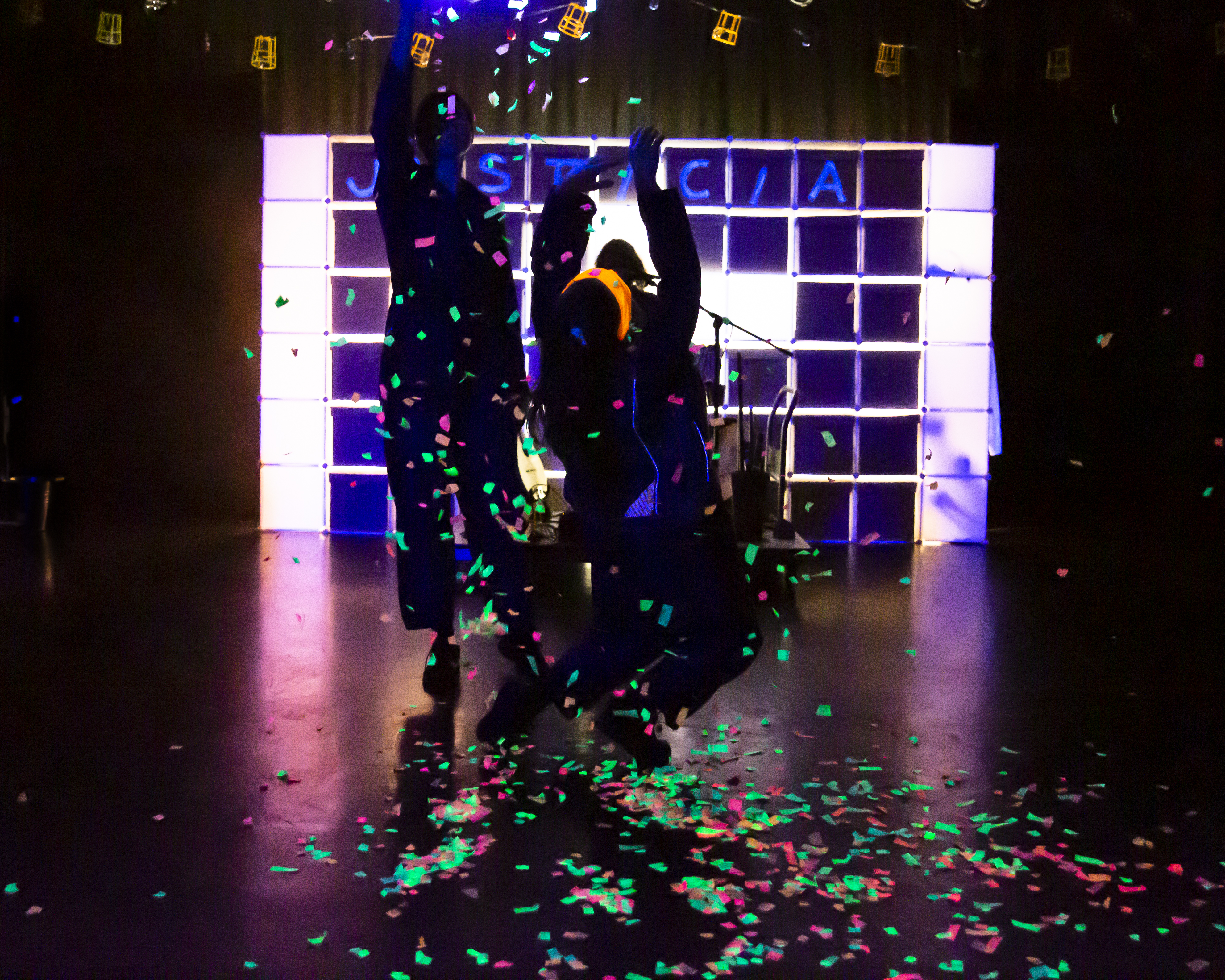 Two people jump amongst colourful confetti, with the word "justicia" visible on the wall behind them
