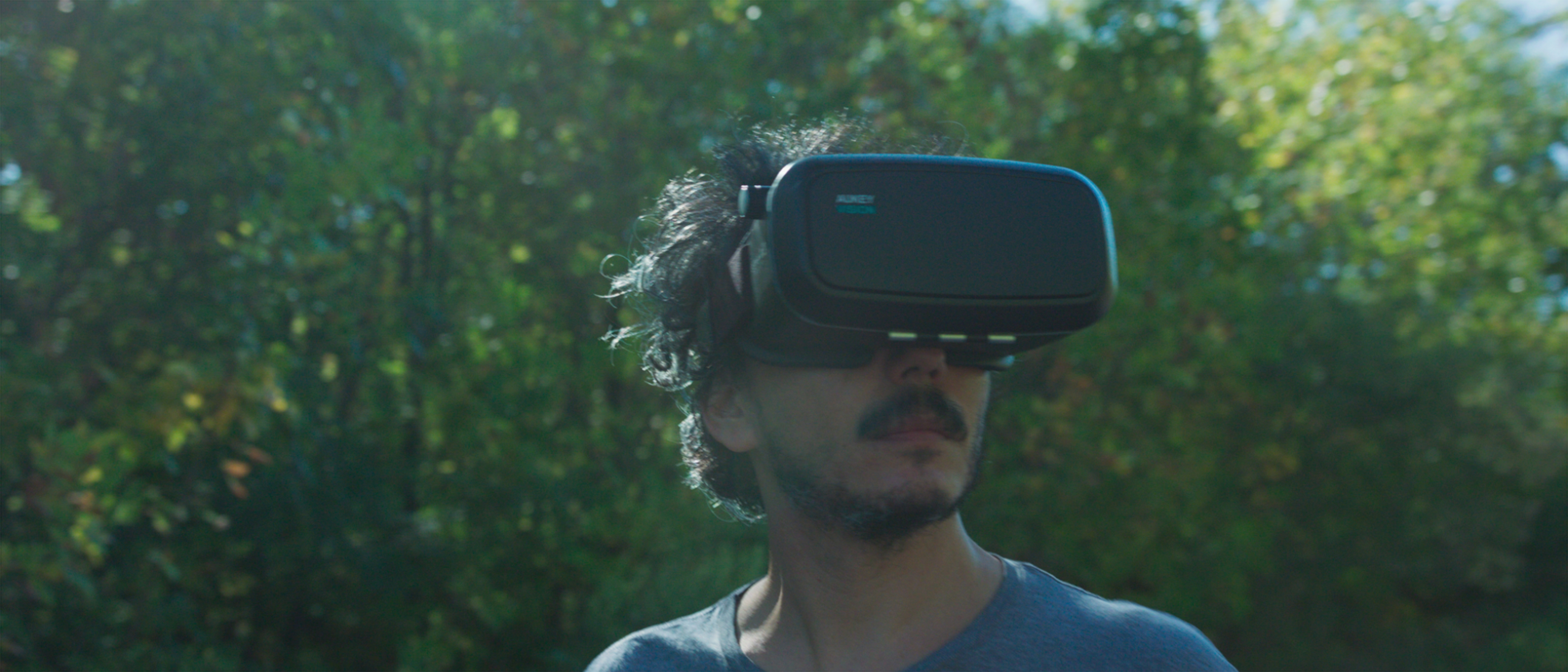 A man in a blue t shirt is stood wearing a VR headset in front of some trees