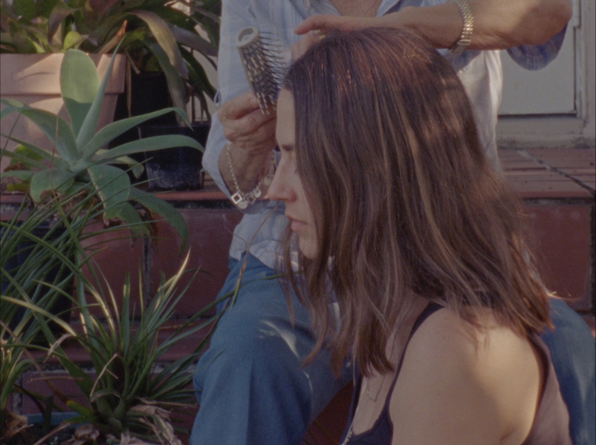 A side view of a woman with long brown hair sat on a step outside a building. Another figure is adjusting her hair and plants are in the background