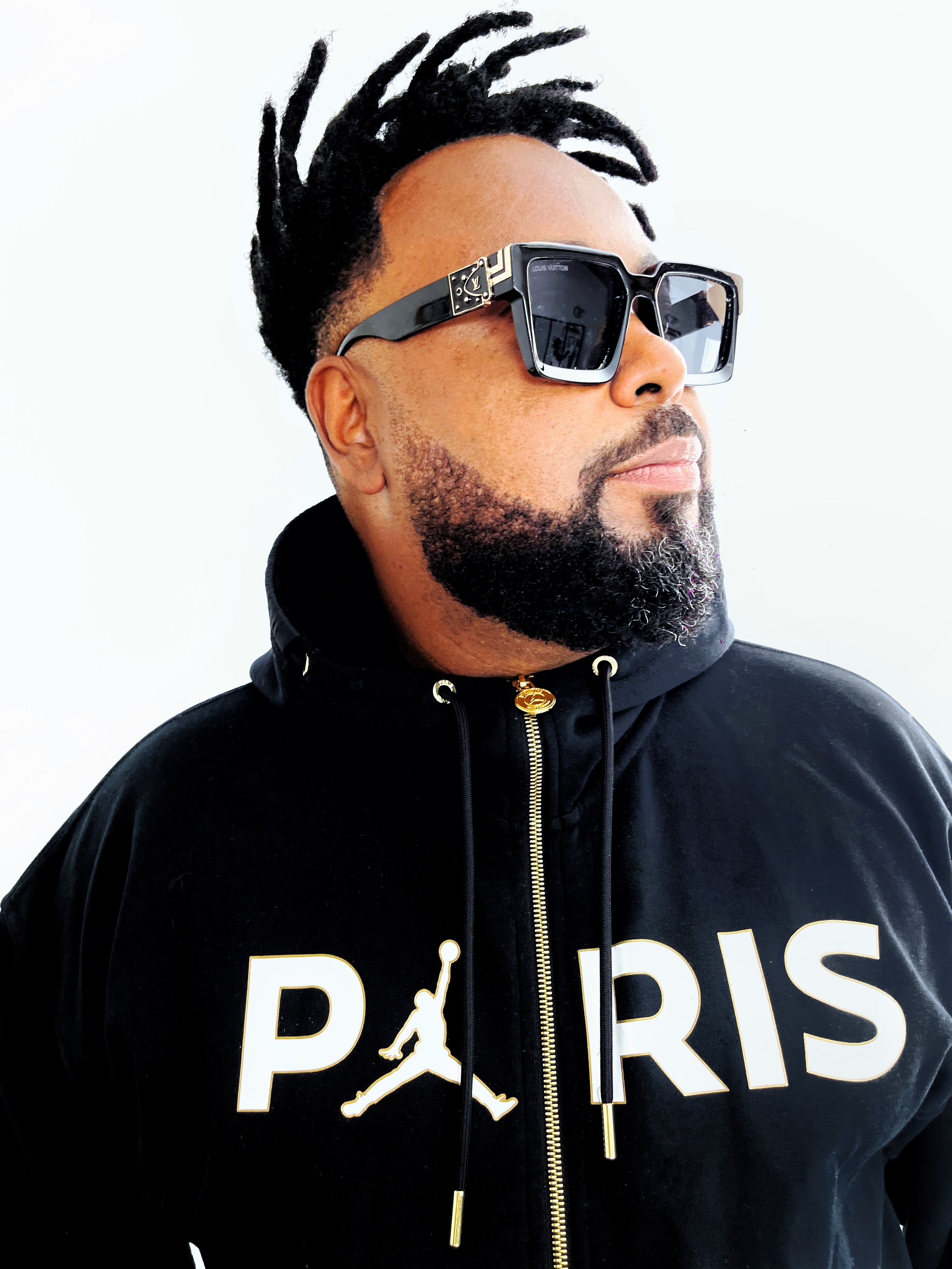 A shot of Angel Yos, wearing a black Nike hoodie with the word PARIS across the chest. He wears square black and gold framed sunglasses, a neatly-trimmed beard, and short dreadlocks. 