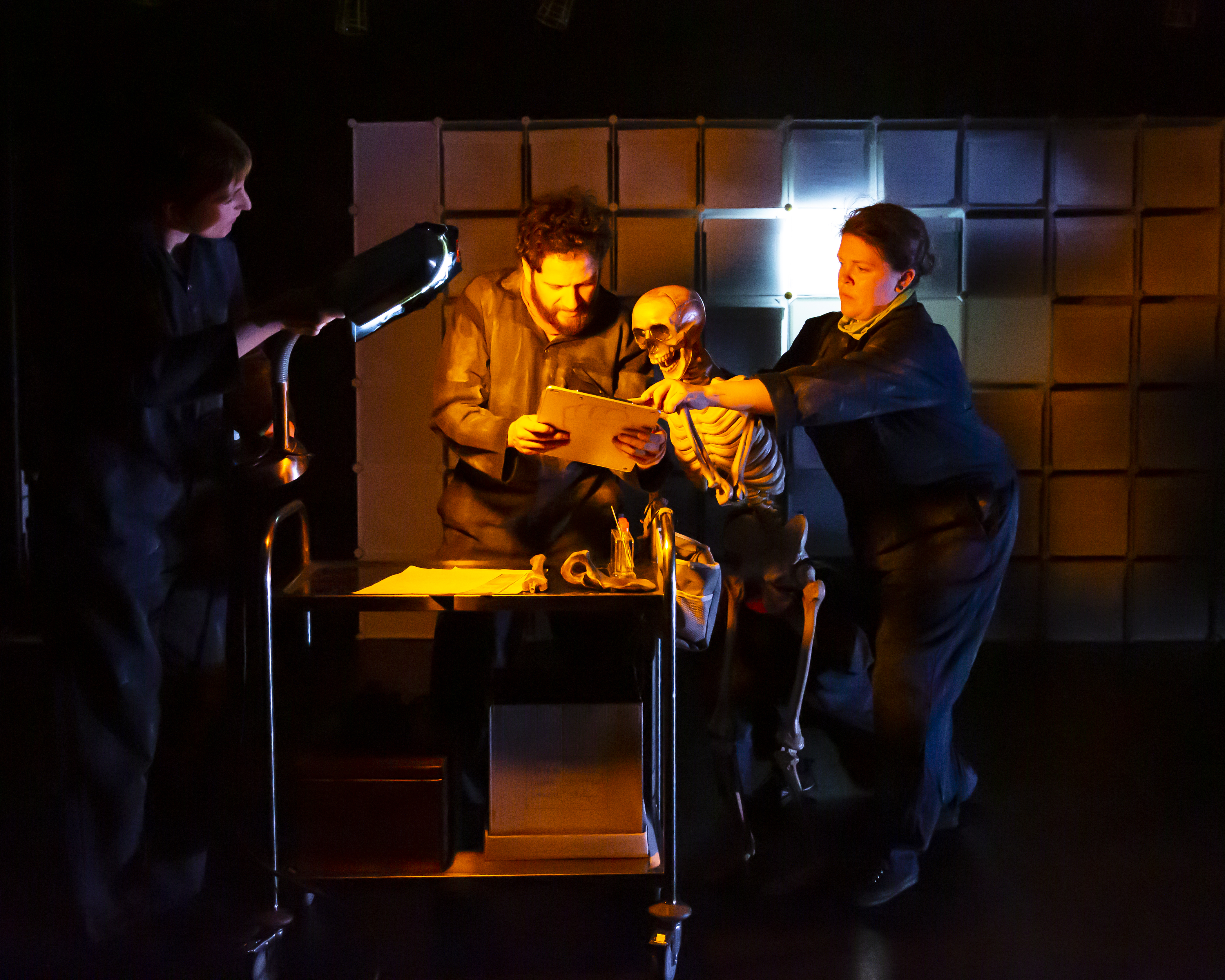 Two people and a skeleton study a clipboard whilst a third person holds a light for them.