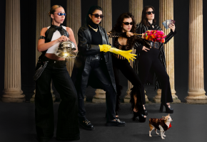 Four Latinx women pose in sunglasses looking mysterious holding a sparkly cloche, a yellow rubber glove, a red feather duster and a handful of passports. A chihuahua stands in a coat in front of them. 