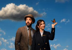 One Mixed race man in a brown tweed jacket and brown bowler hat looks up to a grey cloud in the sky. Next to him, a woman in a navy jacket with gold buttons looks into the camera as an orange butterfly lands on her hand. 